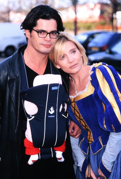 Man holding a baby in a BabyBjörn carrier, with a woman in a blue and yellow costume leaning on him.