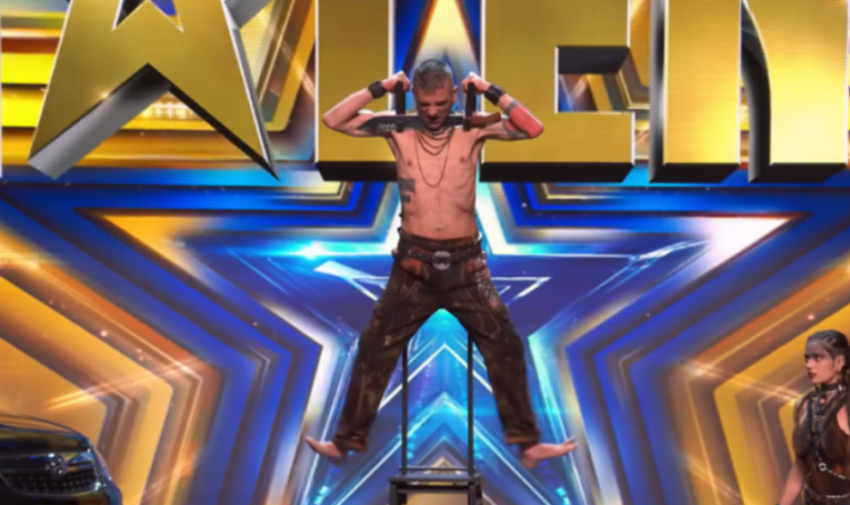 A man with bare chest and dark pants is suspended by shackles around his arms with two swords inserted, on a bright stage.