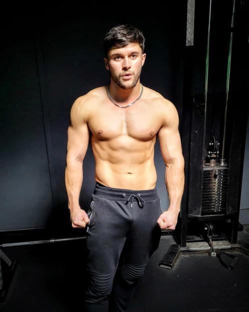 Aedan Duckworth standing shirtless in a gym, wearing dark sweatpants and a silver chain.