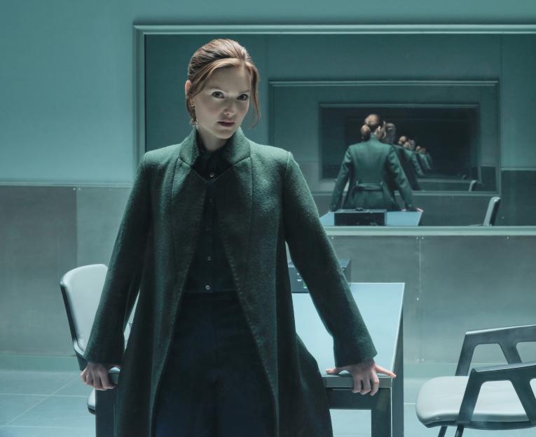 Woman in a dark green coat looking at the camera, with her reflection in a mirror behind her.