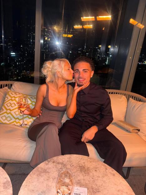 Elsa Rae kissing Marcus John's cheek on a sofa with city lights in the background.