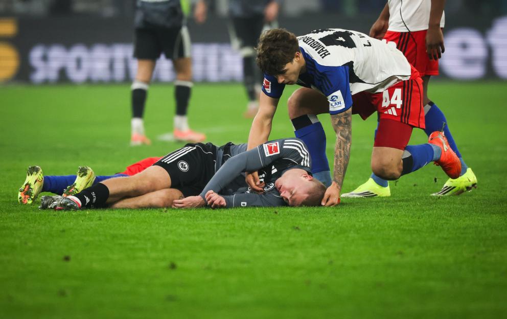 Soccer player in a white and blue jersey helping an injured player in a gray and black jersey on the field.