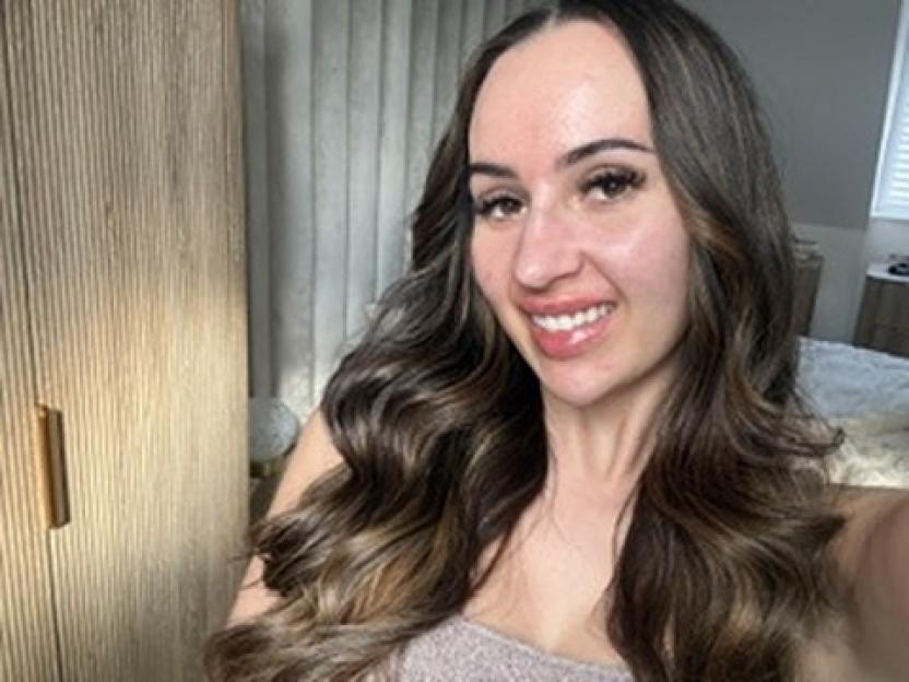 Emily Howdle, a British woman, smiling with her long, wavy brown hair and clear skin.