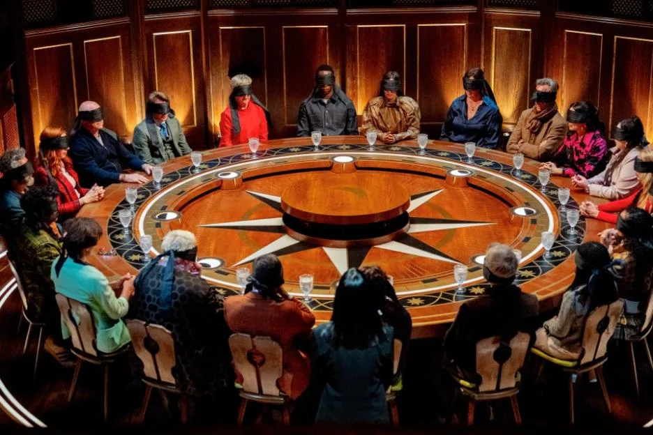 Celebrities wearing blindfolds seated around a round wooden table on The Celebrity Traitors.