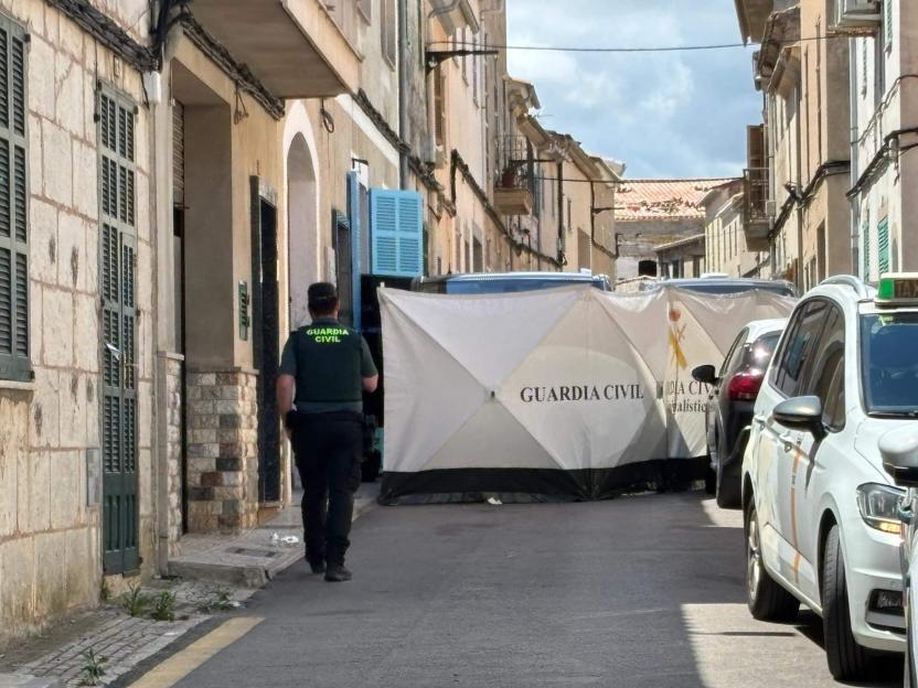A Guardia Civil agent walks past white forensic tents covering an area in a narrow street lined with buildings.