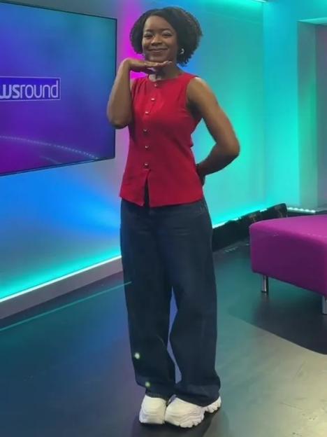 A woman in a red top and jeans posing with her hand under her chin, in front of a monitor displaying "Newsround".