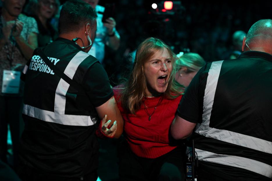 A female protestor in a red shirt shouts as two security personnel escort her from a building.