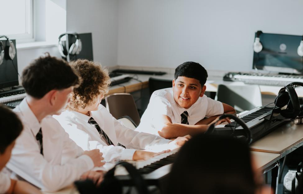 Four high school boys in uniform chat while sitting in a row in a sunny music classroom.