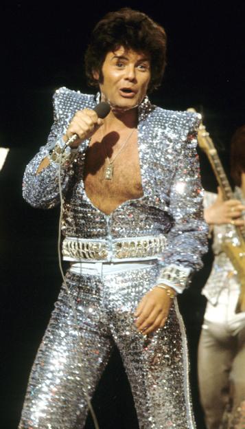 Gary Glitter in a sequined jumpsuit performing with a microphone.