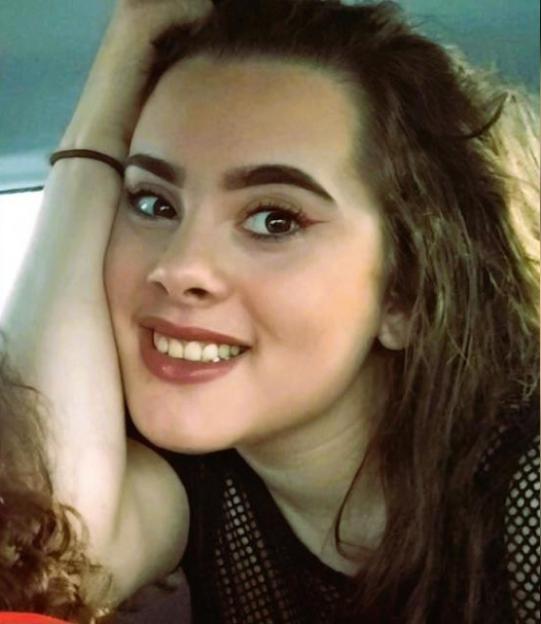 Smiling woman with brown hair, wearing a mesh top, holding her head with her hand.