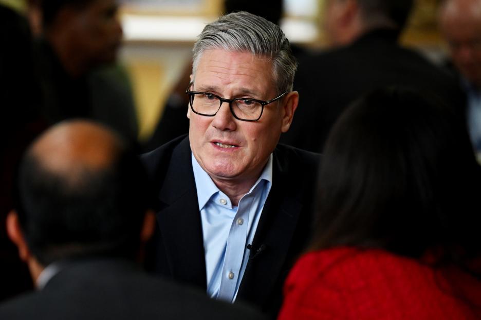 Keir Starmer, wearing glasses and a light blue shirt, speaks to a group of people.