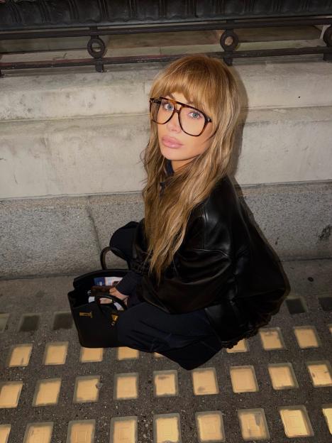 Olivia Attwood in a black leather jacket, dark trousers, and clear square-rimmed glasses, sitting beside a black handbag.