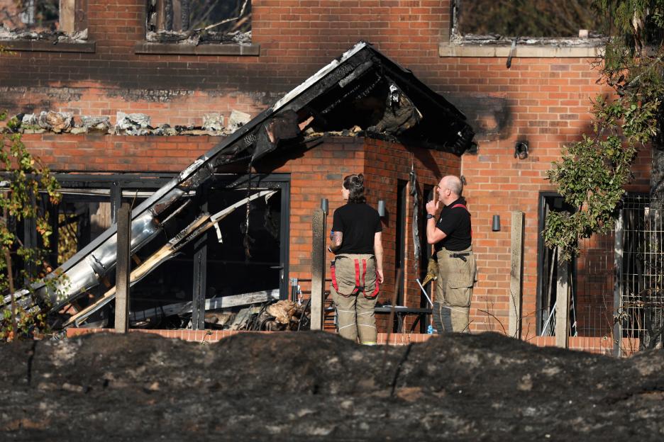 Two firefighters inspect a severely fire-damaged brick house, with a charred roof and blackened windows.
