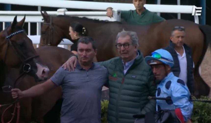 Two men in coats and a jockey in blue stand with horses after a race.