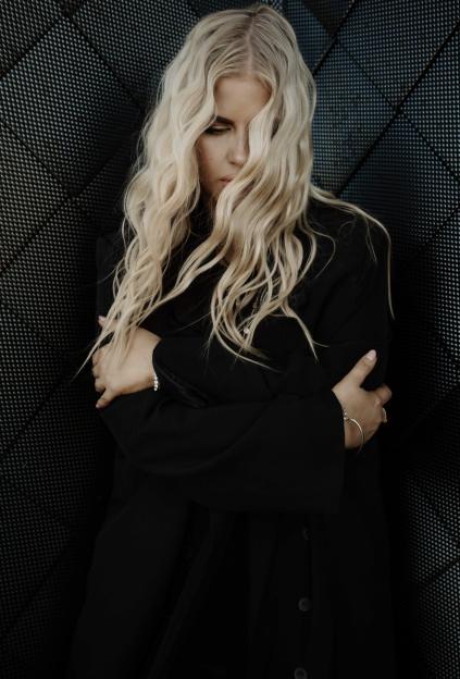 A pop artist, singer, and musician with long blonde hair, wearing a black coat, crosses her arms in front of a dark, textured background.