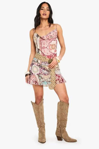 A woman wearing a pink patterned mini dress with a cowl neck, a tan studded belt, and tan cowboy boots.