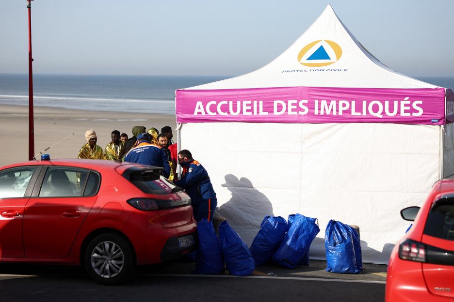 Rescue units aid migrants with thermal blankets next to a tent on a beach.