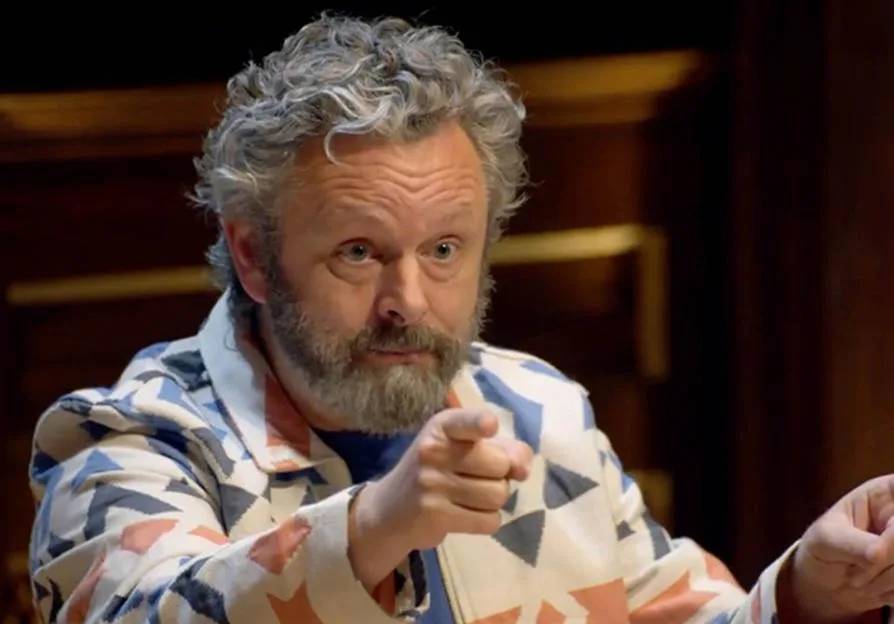Michael Sheen pointing while wearing a patterned jacket.