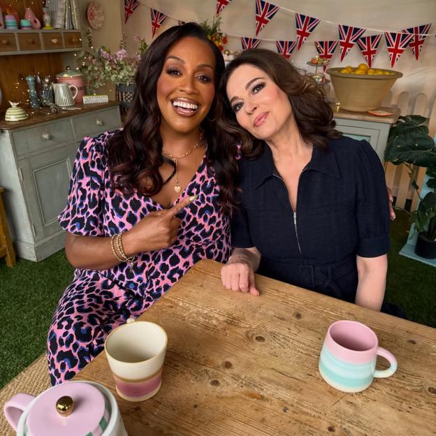 Two women sitting at a wooden table with teacups, one in a pink leopard print dress pointing, and the other in a navy dress.