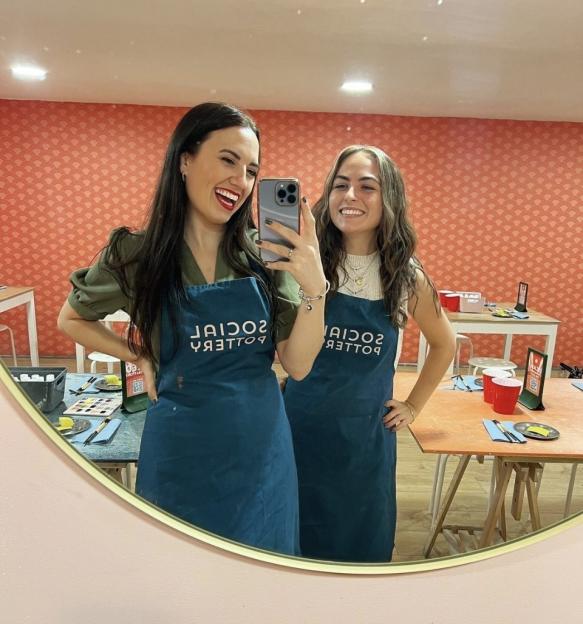 Two young women in aprons reflecting in a mirror, smiling at the camera.