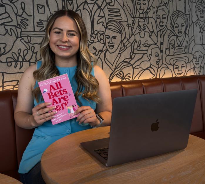 A woman holding the book "All Bets Are Off" by Estelle Maskame, smiling at a table with a laptop.