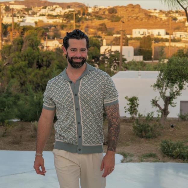 A smiling man with a beard and tattoos on his arms, wearing a patterned button-up shirt and light pants, stands outdoors.