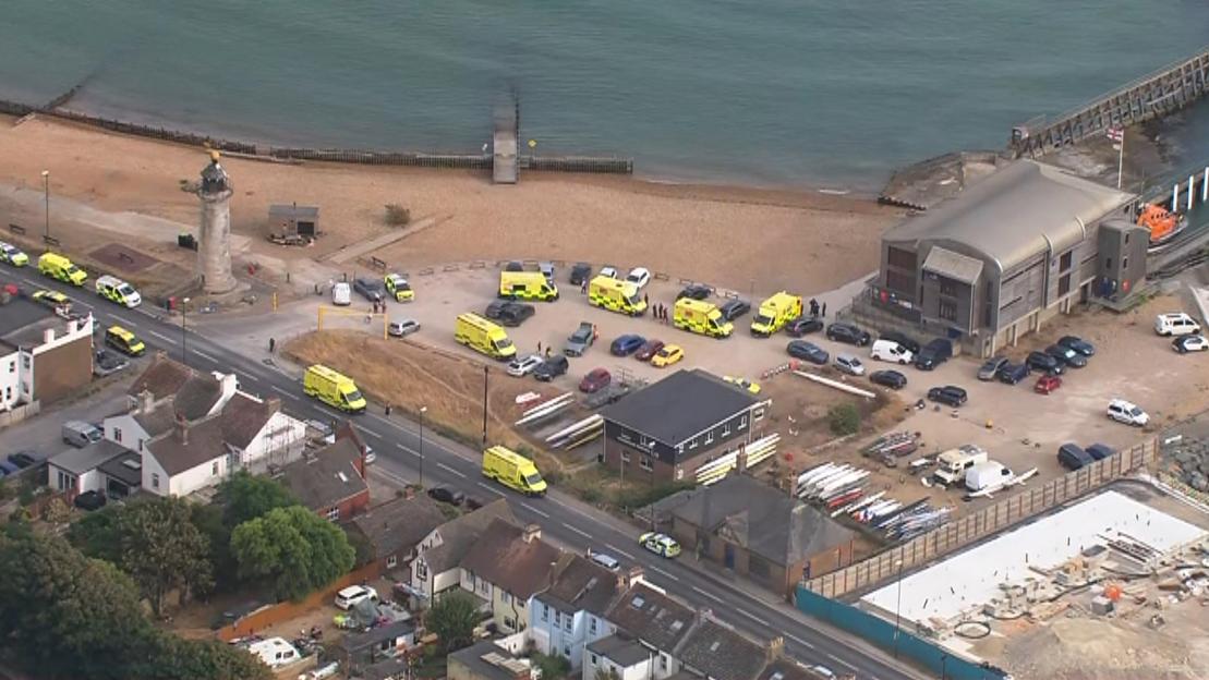 Helicopter footage of Shoreham Harbour showing numerous ambulances and police vehicles parked along the road and beach area.