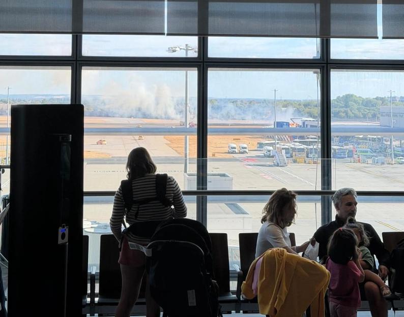 Airport view of smoke from a fire near a runway at Heathrow, with travelers waiting in the foreground.