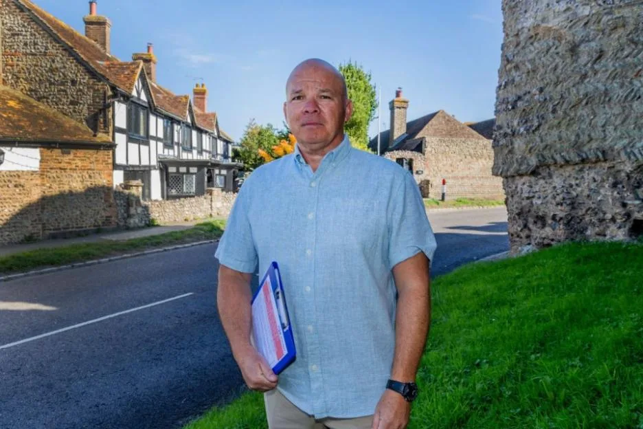 A bald man wearing a light blue shirt holding a clipboard while standing on a street in front of historical houses.