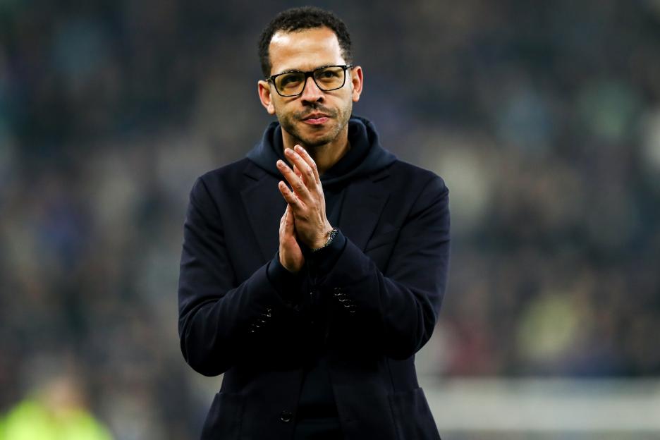 A man in a dark suit and hooded sweatshirt clapping at a soccer match.