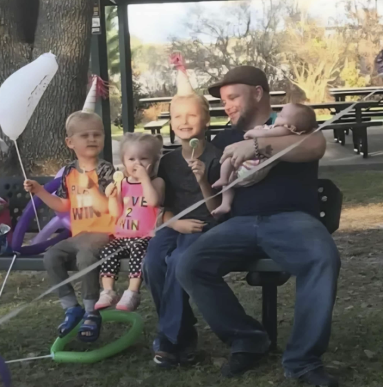 A man and four young children, some wearing party hats and holding balloons, sit together on a bench.