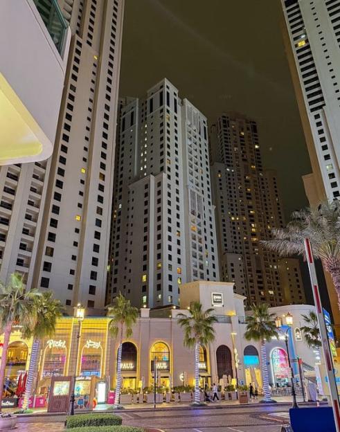 A vibrant street scene at night in Dubai, with tall buildings and storefronts illuminated against a dark sky.