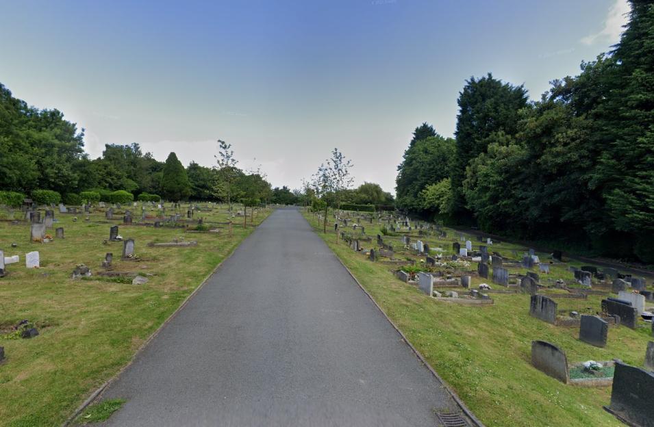 A paved path through a cemetery with gravestones on grassy hillsides under a blue sky.