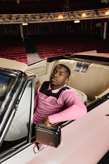 A young man in a pink shirt with black accents sits in a pale pink convertible in a theater with red seats.