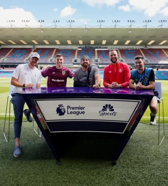 Dude Perfect members pose in front of a Premier League and NBC Sports desk at a stadium.