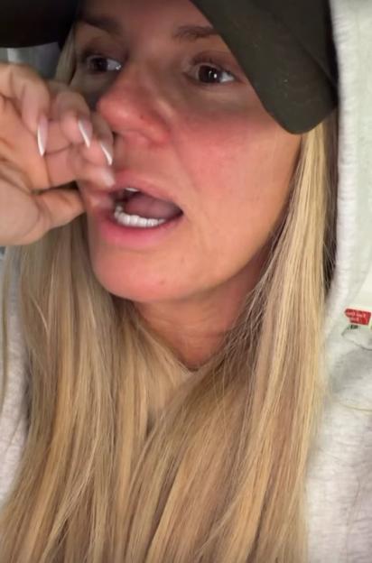 A woman with long blonde hair, wearing a green baseball cap and a white hooded sweatshirt, holds her hand near her mouth as she speaks.