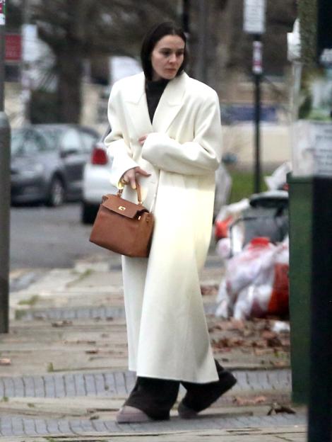 A guest leaving Gordon Ramsay's house, wearing a long white coat, dark turtleneck, and carrying a brown handbag.