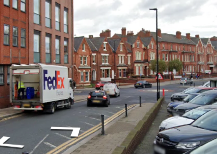 A FedEx truck with its rear door open, and a person in a safety vest working in the back, is parked on a street with other cars and brick buildings.