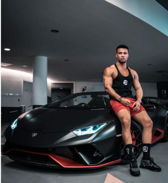A muscular man in a black tank top and red shorts sitting on the hood of a black Lamborghini.