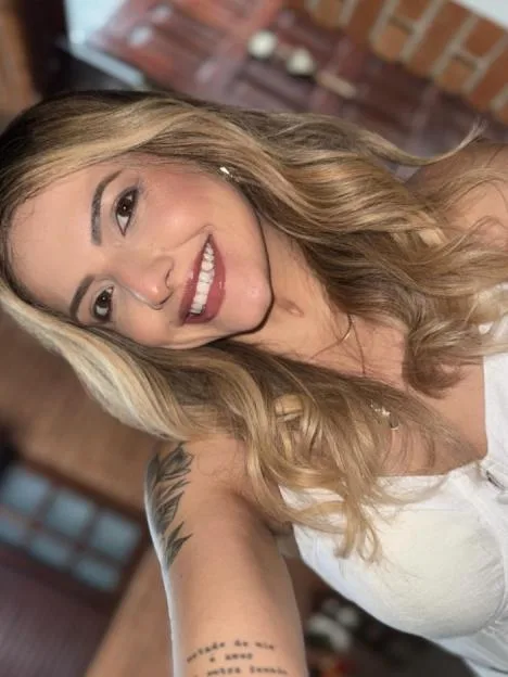 A woman with long blonde hair and a white top smiles at the camera, with a tattoo on her arm.