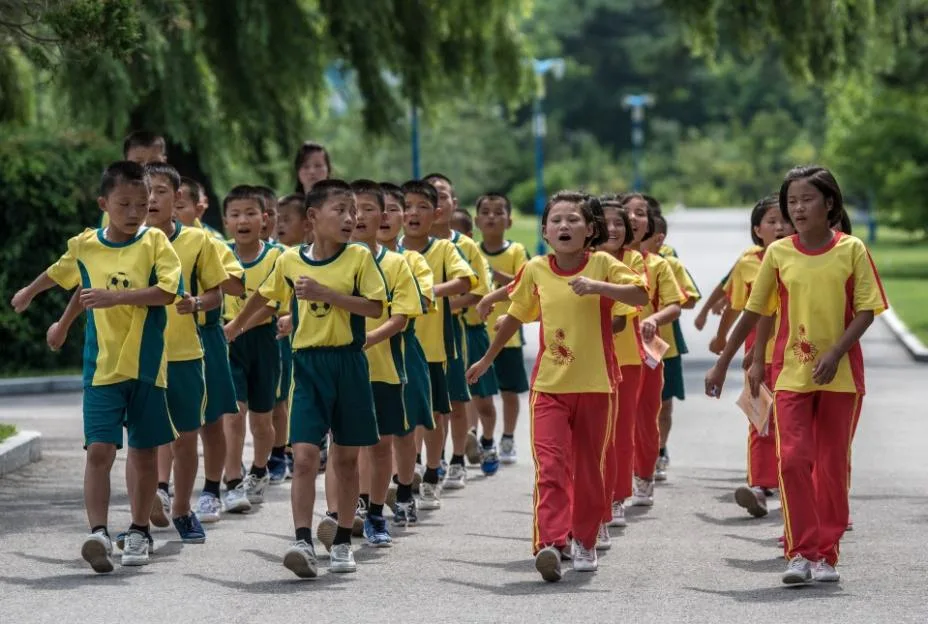A large group of North Korean children in matching yellow and green or red uniforms marching down a paved path outdoors.