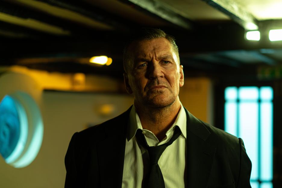 Craig Fairbrass, an actor with short gray hair and a stern expression, wearing a black suit jacket and a loosened black tie, looks directly at the camera.
