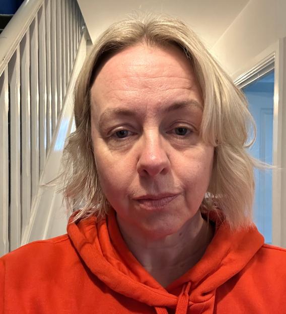 A blonde woman in an orange hoodie looking directly at the camera.