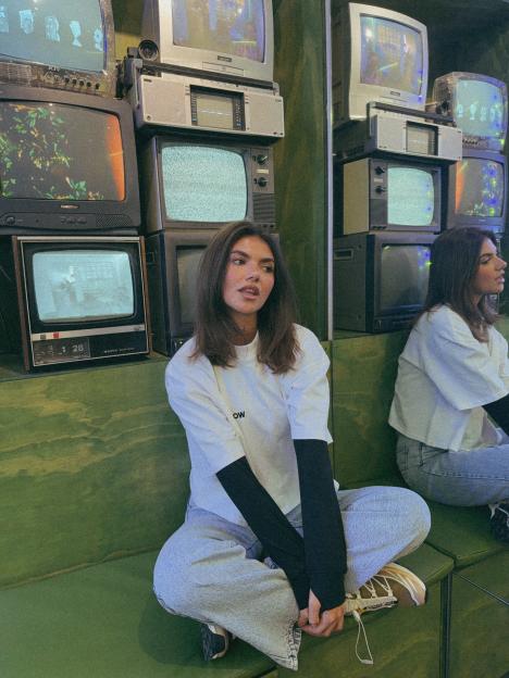 Samie Elishi sitting on a green couch in front of a wall of vintage televisions.