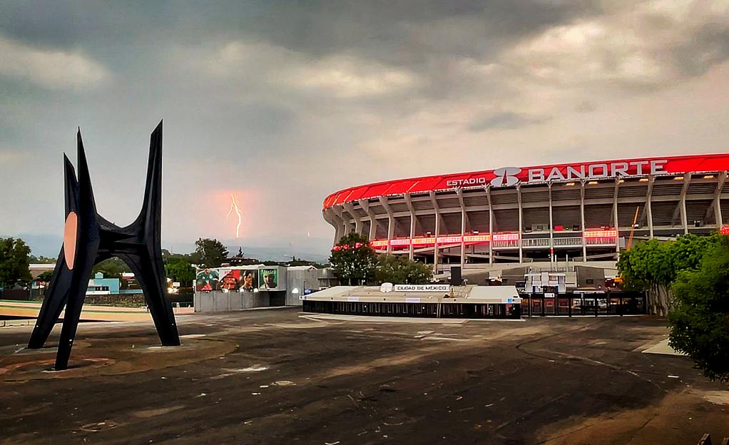 Estadio Banorte in Mexico City under a cloudy sky with lightning in the distance.