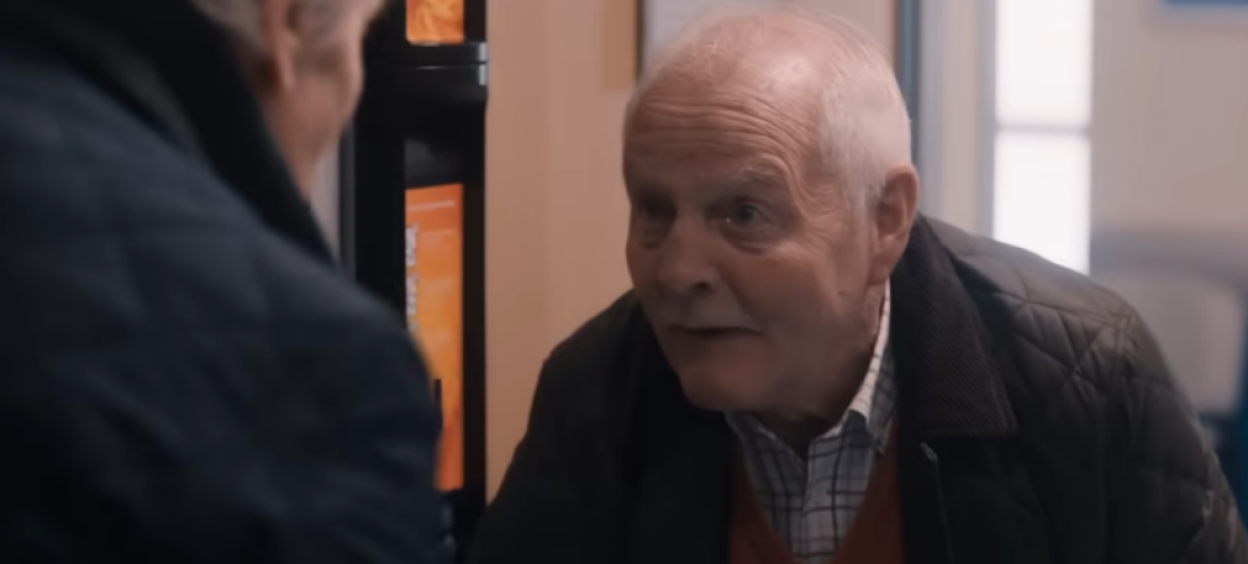 An older white man with white hair, wearing a patterned shirt, a red sweater, and a dark quilted jacket, looking at another person off-screen.