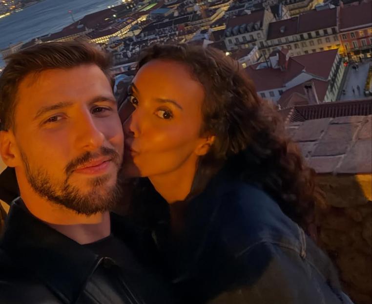 Maya Jama kissing Ruben Dias on the cheek with a city skyline at dusk in the background.