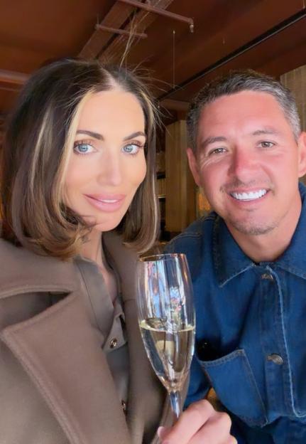 A woman in a brown coat holding a glass of champagne, and a man in a denim jacket, smile at the camera.