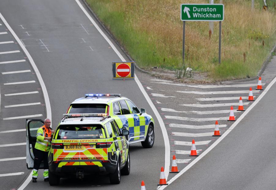 Traffic officer exiting a "Highways England" vehicle next to a police car, with an emergency service sign, and traffic cones blocking a road.