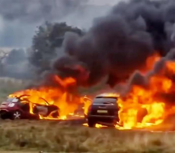 Two cars engulfed in flames in a field, with dark smoke rising into the sky.